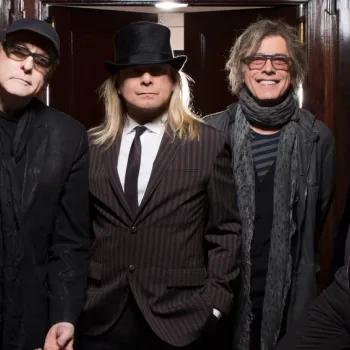 Four men stand side by side on a staircase, wearing dark clothing, scarves, and jackets; one wears a hat, and three have long hair.