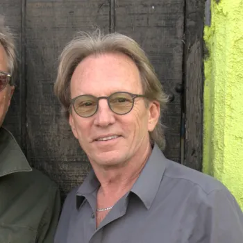 Two older men with light hair and glasses stand side by side, smiling, in front of a weathered wooden door and a brightly painted wall.