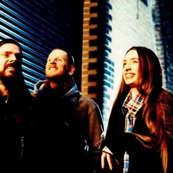 Four people stand outside near a brick wall and metal gate, dressed in casual jackets, looking slightly upward with neutral expressions.