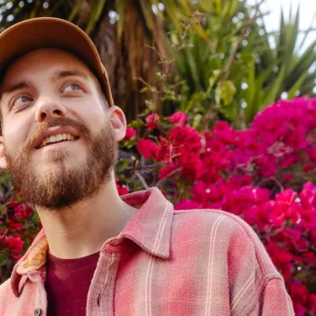 A bearded man in a cap and pink jacket stands outdoors, smiling, with bright pink flowers and green plants in the background.