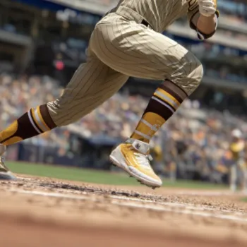A baseball player in a brown and yellow uniform runs to first base during a game, with a blurred crowd in the background.