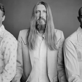 Three men pose together against a plain backdrop, each wearing light-colored, patterned shirts; one has long hair, one has short hair, and one is bald.