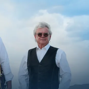 Two older men wearing sunglasses, white shirts, and black vests stand outdoors in front of a cloudy sky. One holds a guitar by his side.
