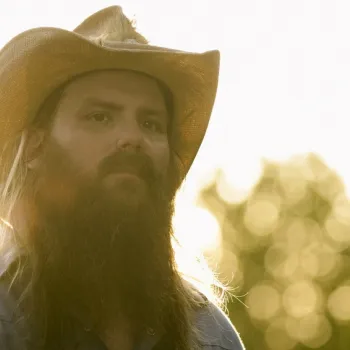 A man with a long beard and mustache wears a cowboy hat and denim shirt, standing outdoors with sunlight and blurred trees in the background.