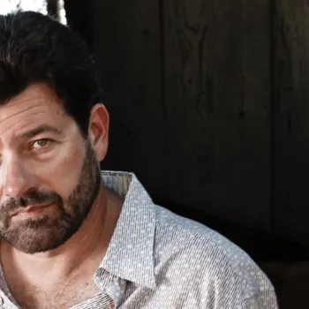 A man with dark hair and a beard, wearing a light-colored button-up shirt, looks at the camera while standing in a shaded area.