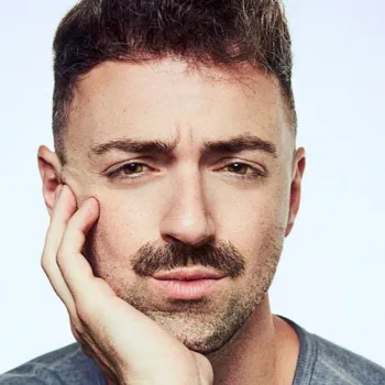 A man with short brown hair and a mustache rests his face on his hand, looking directly at the camera against a plain light background.