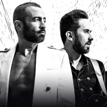 Two men stand side by side; one is shirtless under a white jacket, the other wears a dark vest and holds an electric guitar. Abstract light rays emanate behind them.