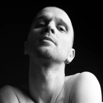 A person with a shaved head looks slightly upward with a neutral expression against a dark background. Their shoulders are visible and one earring can be seen.