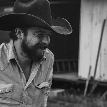 A bearded man in a cowboy hat and denim shirt stands outdoors near a building, looking to the side and smiling. The image is in black and white.