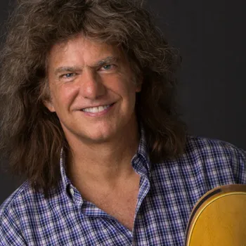 A man with long curly hair wearing a purple checkered shirt smiles while holding a yellow guitar against a dark background.