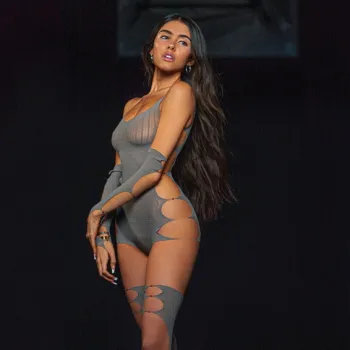 A woman with long dark hair wears a fitted, asymmetrical gray bodysuit with large cut-outs and matching thigh-high stockings, standing against a plain dark background.