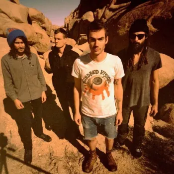 Four men stand outdoors among large rocks, facing the camera. One wears a white t-shirt and shorts; the others wear darker clothing and hats. Shadows and sunlight suggest a desert setting.