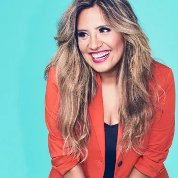 A woman with long wavy hair wearing a bright orange blazer over a black top smiles against a light blue background.