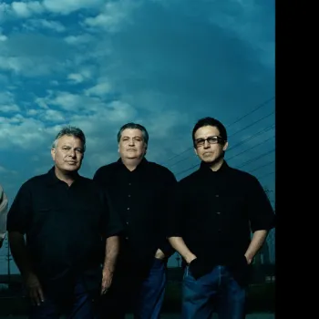 Five men stand outdoors under a cloudy sky, wearing dark clothing. Four are wearing glasses or sunglasses, and each has their hands in their pockets.