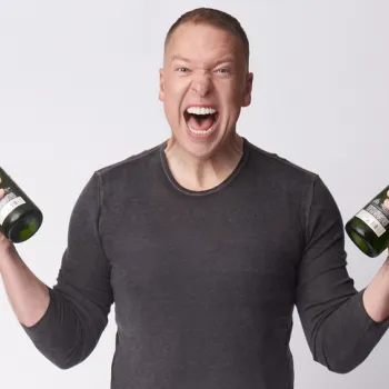 A man in a gray shirt holds a bottle of champagne in each hand and yells with excitement against a plain white background.