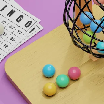A bingo game setup with bingo cards, a white ball on one card, colorful balls on a wooden surface, and a metal cage for drawing balls on a purple background.