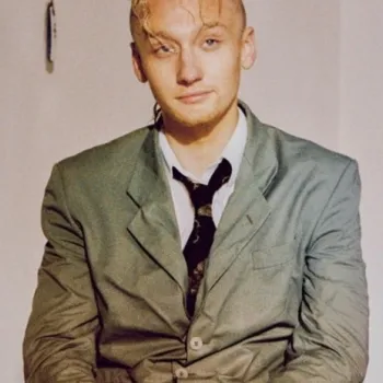 A person with short, wet-looking hair sits indoors wearing a crumpled green suit, white shirt, and black patterned tie, looking at the camera with a neutral expression.