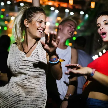 Three young adults dance energetically in a club or concert setting with colorful lights in the background.