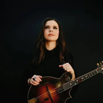 A woman with long brown hair stands against a dark background, holding a mandolin and looking slightly upward.