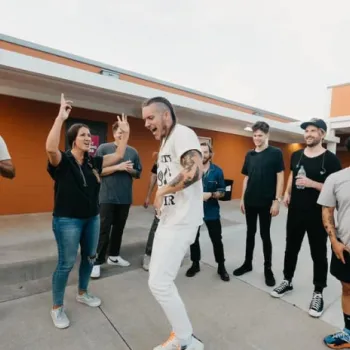 A group of people stands and talks outside a building; one man in white is dancing while others watch and smile.