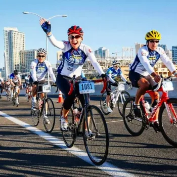 A group of cyclists in helmets and event jerseys ride on a city street during a race; one cyclist waves and smiles at the camera. Tall buildings are visible in the background.