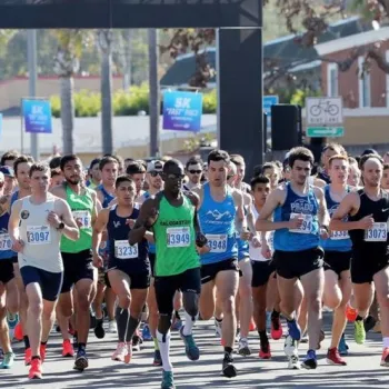 A large group of runners begins a road race, wearing numbered bibs and athletic clothes, with spectators and race banners visible in the background.