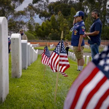 A boy in a Cub Scout uniform salutes a gravestone in a cemetery, accompanied by an adult, with several American flags placed by headstones.