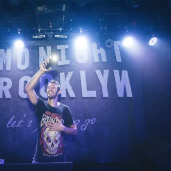A man stands on stage in front of a sign reading "Emo Night Brooklyn," holding up a phone and smiling under stage lights.