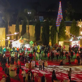 Crowd of people walk through a festive outdoor plaza at night, with colorful lights, decorated trees, and holiday market booths visible.