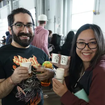 Two people are smiling and holding cups at an indoor event with other people in the background. The woman is holding a stack of cups; the man holds several drinks.