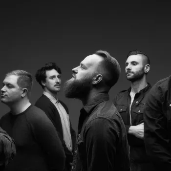 Six men standing and facing different directions against a dark, plain background. All wear casual clothing; some have beards and glasses. The setting appears to be a studio.