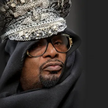A person wearing oversized glasses, a sparkly embellished hat with jewels, and a black cloak, looking slightly to the side against a dark background.