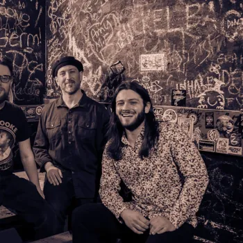 Four men pose together in a dimly lit room with graffiti-covered walls and a bench, smiling at the camera.