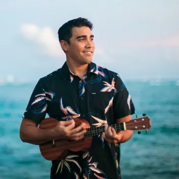 A man wearing a black floral shirt stands by the ocean, smiling and playing a ukulele with mountains visible in the distance.