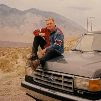 A person in a denim jacket sits on the hood of a black Ford 4x4 truck parked in a dry, mountainous landscape.