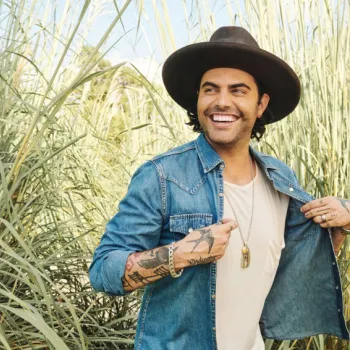 A smiling man with tattoos, wearing a wide-brimmed hat and denim shirt over a white t-shirt, stands outdoors in tall grass.