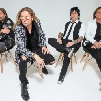 Four men sit on wooden chairs against a plain white background, dressed in stylish black and white clothing, looking toward the camera.