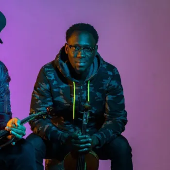 Two men sit side by side holding violins, one in a red jacket and cap, the other in a camo hoodie, against a pink and blue lit background.