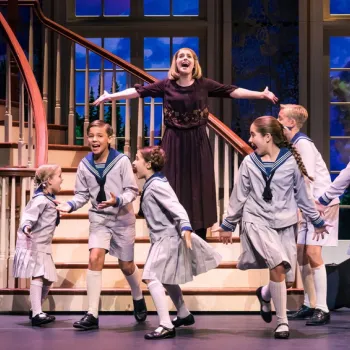 A woman sings joyfully on stage with seven children in sailor-style outfits in front of a staircase and large windows during a theatrical performance.