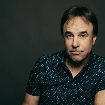 A man with short brown hair wearing a navy blue shirt with white dots sits against a dark background, looking directly at the camera with a neutral expression.