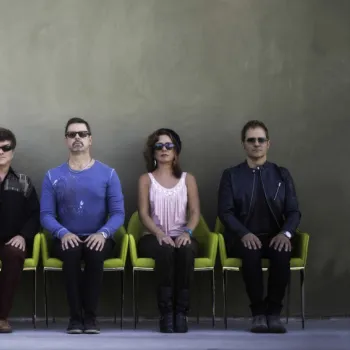Six people in sunglasses sit in a row of green chairs against a plain wall, with one person on the far left facing sideways.