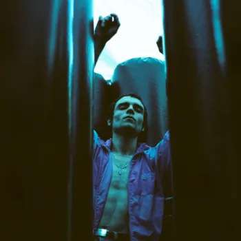 A person with a bare chest and open shirt stands with arms raised between two dark vertical structures, illuminated by blue-green light.