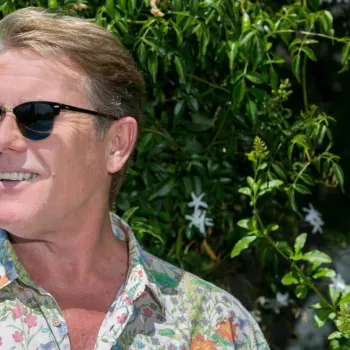A man wearing sunglasses and a floral shirt stands in front of green foliage and blooming flowers, smiling.