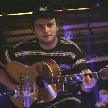 A person wearing a striped shirt and cap plays an acoustic guitar in a music studio, seated in front of a piano and a microphone.