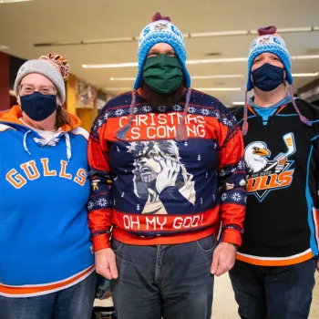Three people wearing face masks, beanies, and hockey jerseys pose indoors; the middle person’s sweater reads “Christmas is coming OH MY GOD!” with a surprised face graphic.