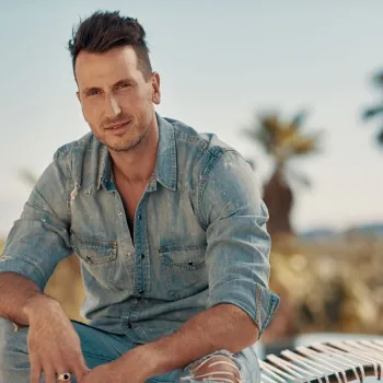 A man in a light denim shirt and ripped jeans sits outdoors on a hammock with palm trees in the background.