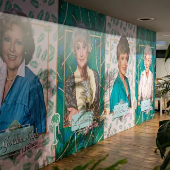 Wall mural featuring large portraits of the four main characters from “The Golden Girls,” each labeled with their name, in a room with plants and wooden flooring.