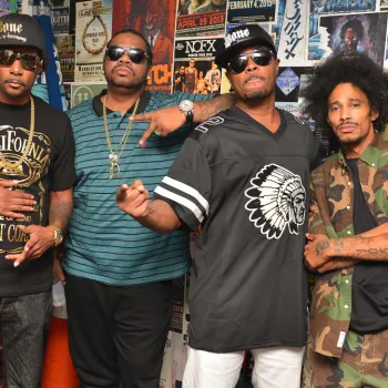 Four men pose in front of a wall covered with colorful posters and flyers; each wears casual streetwear, including sunglasses, hats, and jewelry.