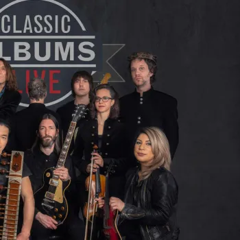 A group of ten musicians pose with instruments in front of a sign that reads "Classic Albums Live" on a dark background.