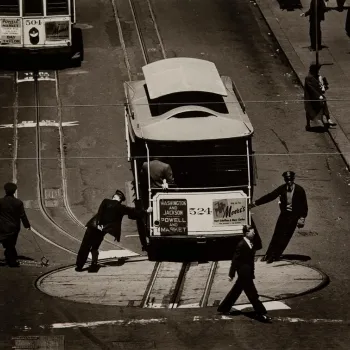 A streetcar is being manually turned on a rotating platform by uniformed workers and men in suits, while pedestrians walk nearby on a city street.